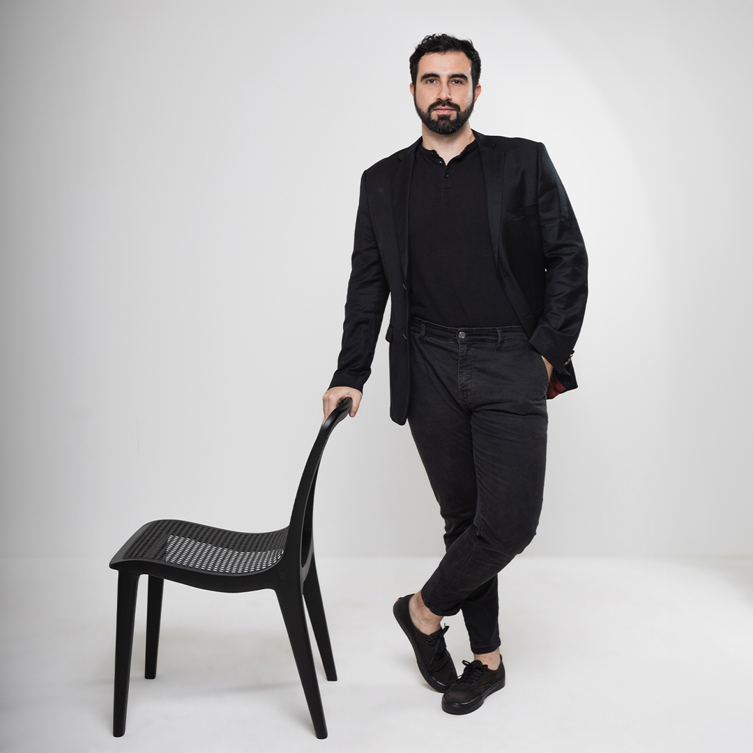 Look profissional em estúdio com composição minimalista em preto total. Fotografia em fundo branco com cadeira moderna preta de design contemporâneo com assento vazado em padrão geométrico. Vestuário elegante all black composto por blazer, camiseta e calça slim, complementado por tênis casual. Pose descontraída apoiada na cadeira moderna de linhas clean e estrutura leve.