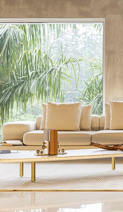 Sala de estar luxuosa e minimalista com sofás modulares em tom bege, decorada com mesa de centro dourada sobre tapete claro. Ambiente contemporâneo com grandes janelas panorâmicas emolduradas por palmeiras tropicais, criando uma atmosfera zen e sofisticada. Modelo veste conjunto branco relaxado em ambiente clean e iluminado com decoração moderna.