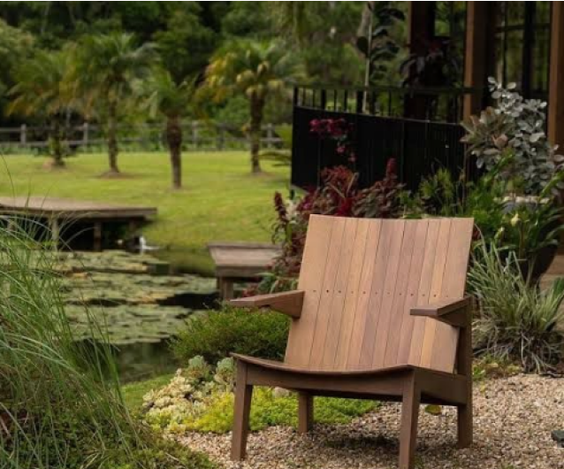 Poltrona de madeira estilo contemporâneo em área externa com design ripado em tom natural claro, braços em madeira escura e estrutura robusta. Posicionada em jardim paisagístico com lago ornamental, sobre caminho de pedriscos, cercada por gramado verde e vegetação variada incluindo capins ornamentais e flores vermelhas. Ambiente zen com paisagismo natural e área de contemplação próxima a espelho d'água.