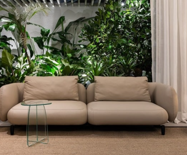 Sofá moderno em tom bege neutro com design minimalista, acompanhado de mesa lateral delicada em metal verde. Ambiente decorado com exuberante jardim vertical de plantas tropicais ao fundo, incluindo palmeiras e folhagens diversas. Decoração contemporânea que integra natureza e mobiliário sobre carpete em tom neutro, complementada por cortina branca lateral.