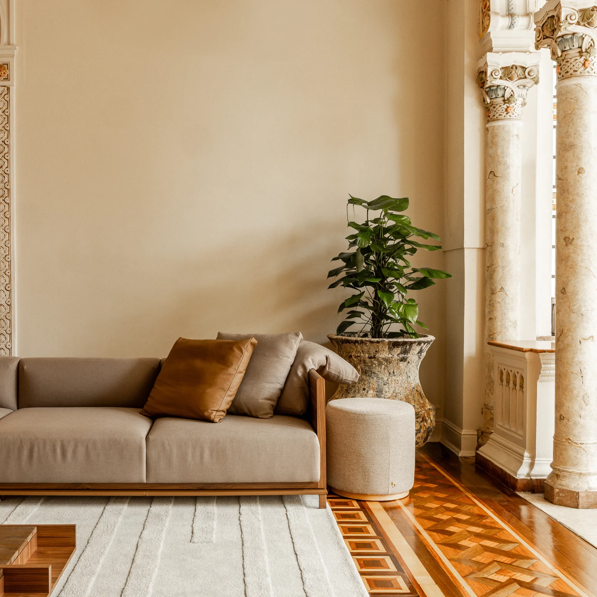 Sala de estar elegante em estilo clássico europeu com sofá moderno em tom cinza e acabamento em madeira. Ambiente luxuoso com teto ornamentado, lustre de cristal dourado e arcos decorativos em estilo Art Nouveau. Piso em marchetaria de madeira, tapete claro texturizado e almofadas em tons terrosos complementam a decoração. Destaque para porta em arco com detalhes ornamentados que conecta a ambientes adjacentes em tons claros e iluminação aconchegante.