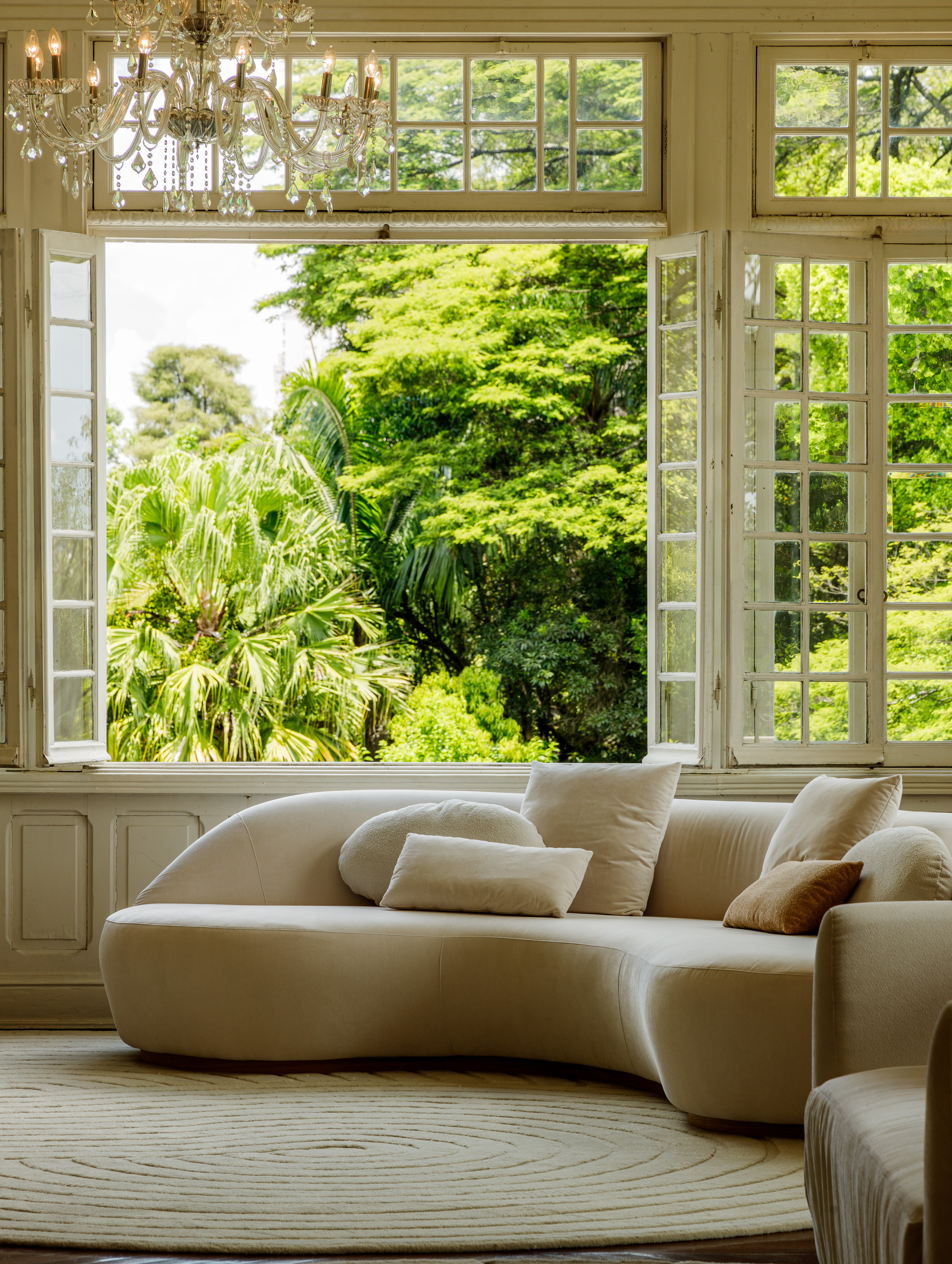 Sala de estar elegante com sofá curvilíneo em veludo bege claro, decorado com almofadas em tons neutros, posicionado sob janelas francesas amplas com vista para jardim tropical exuberante. Ambiente sofisticado iluminado por lustre de cristal clássico, piso em madeira clara e tapete circular com padrão orgânico em tons claros. Vista panorâmica enquadra palmeiras e vegetação verde vibrante, criando integração perfeita entre interior luxuoso e natureza exterior.
